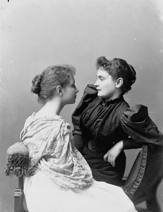 Hellen Keller and Anne Sullivan, wearing Victorian clothing and hairstyles, are seated looking into each others' faces